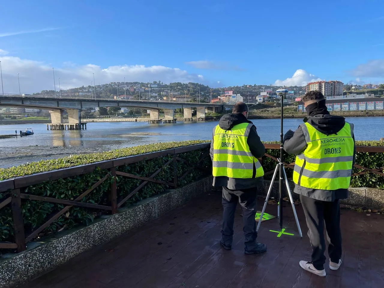 Two Aeromedia operators in hi-vis vests with a GNSS station beside an estuary at low tide, with a bridge in the background