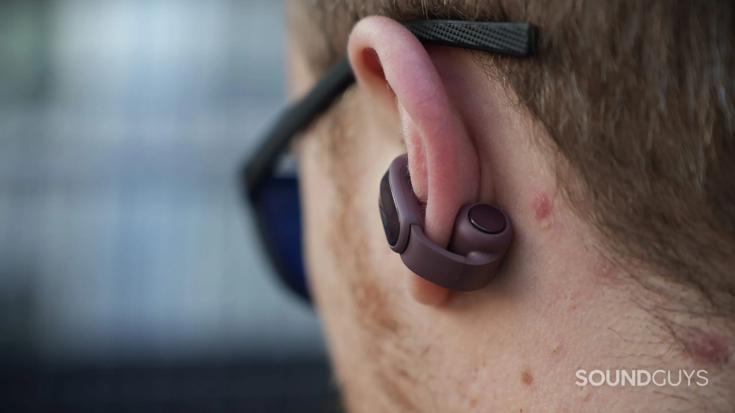 Primer plano trasero de un auricular Bose Ultra Open 2 rodeando la oreja bajo unas gafas de sol