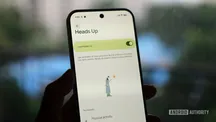 Una mano sostiene un móvil Pixel con la pantalla de ajustes de Heads Up y el interruptor de activación encendido, sobre un fondo exterior desenfocado