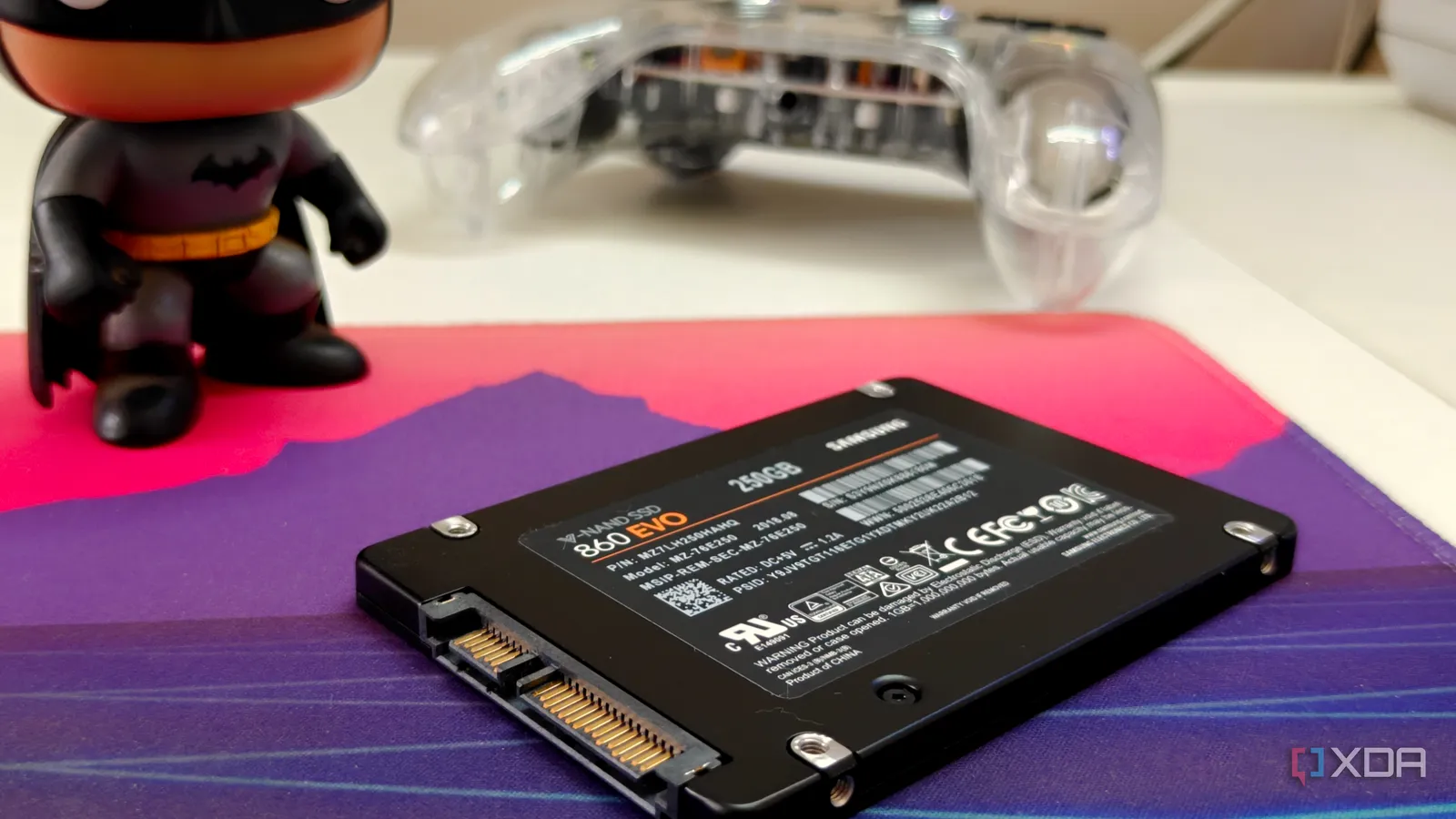 Un SSD SATA junto a un teclado y un mando de consola sobre una alfombrilla