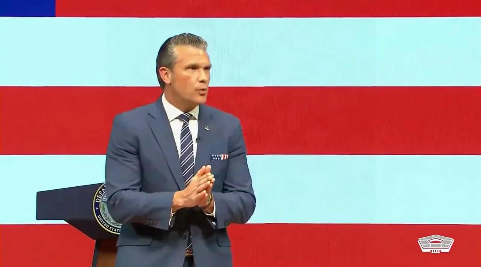 Pete Hegseth, in a navy suit, gesturing with his hands in front of red, white and blue stripes and a lectern bearing the Department of War seal