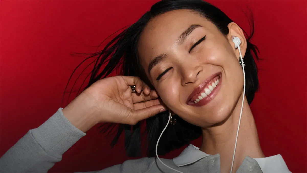 Woman smiling as she listens to music with the white Bose Noise Cancelling Wired Earbuds on a red background