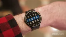 Wrist wearing the Pebble Round 2: black round face with white digits and dark strap over a checkered sleeve