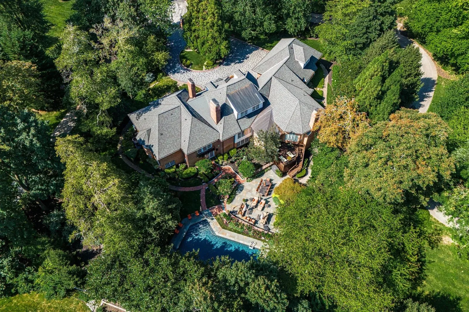 Top-down view of a house with a grey roof, brick patio and swimming pool surrounded by trees, shot from a drone