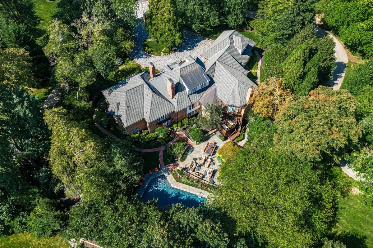 Vista cenital de una casa con tejado gris, patio de ladrillo y piscina rodeada de árboles, tomada con dron