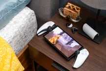 The PlayStation Portal on a bedside table, next to a pair of earbuds, streaming God of War: Ragnarök from a PS5