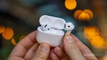 Un AirPod sujeto junto a su caja de carga abierta, con luces cálidas de fondo