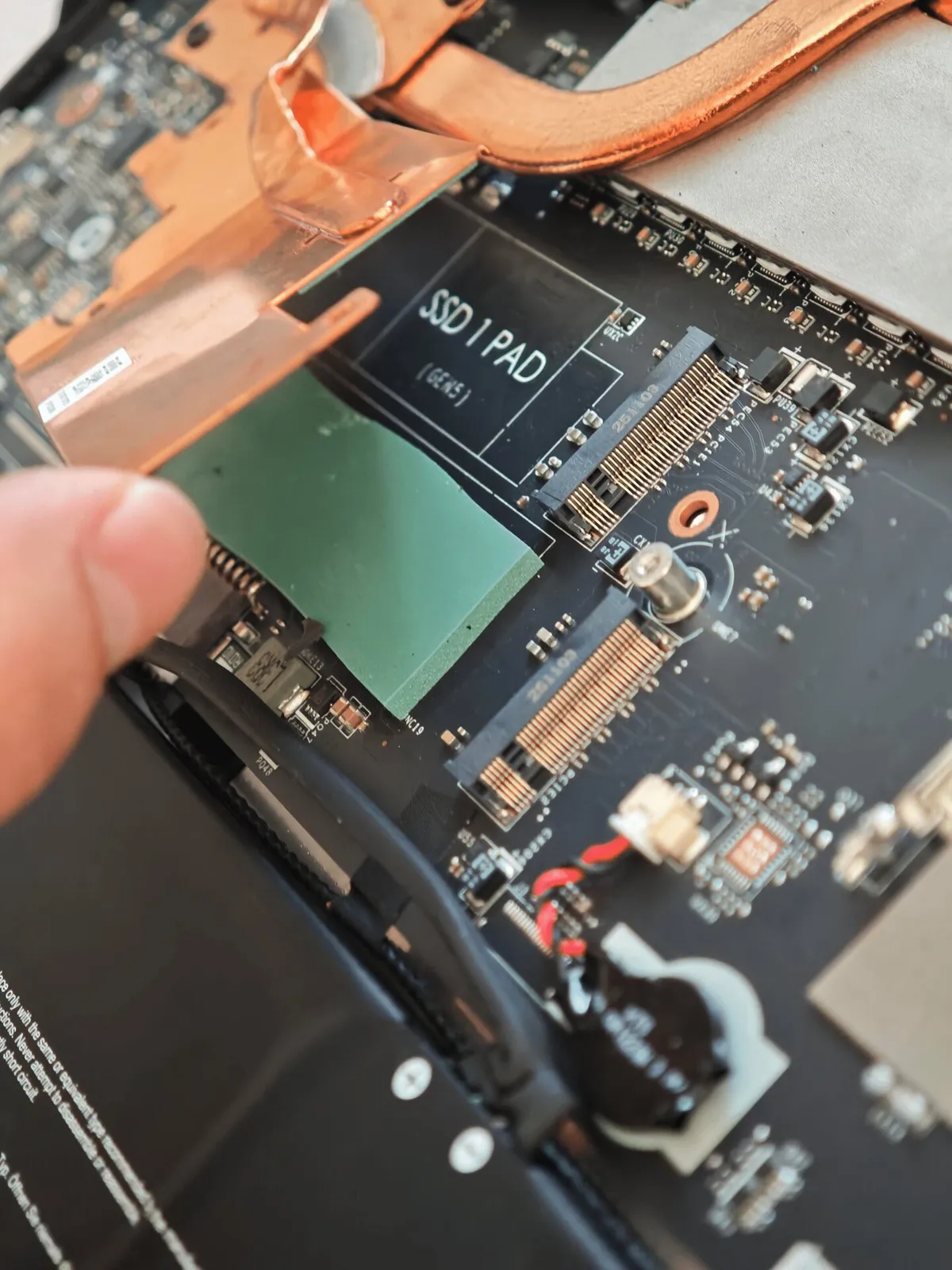Mano levantando el disipador de cobre situado sobre el zócalo «SSD 1 PAD» de un portátil gaming