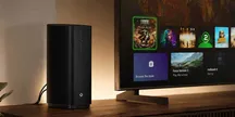 Xbox Series X console standing vertically on a wooden cabinet, next to a TV displaying the main Xbox menu