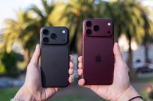 Dos iPhone de última generación sostenidos con una mano, uno en negro y otro en granate, con palmeras al fondo