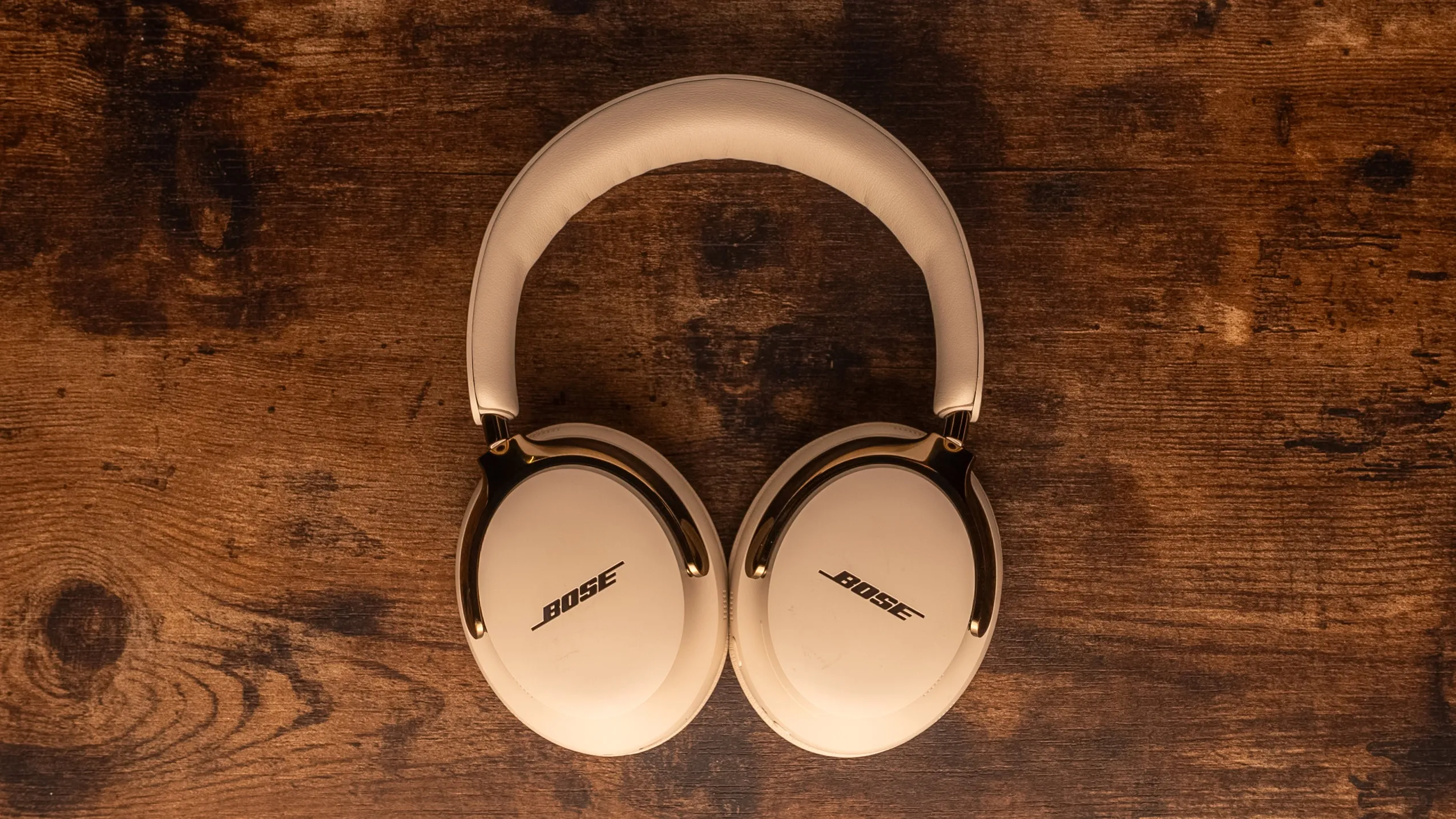 Bose QuietComfort Ultra (2nd Gen) headphones in a light color with gold accents, resting on a wooden table