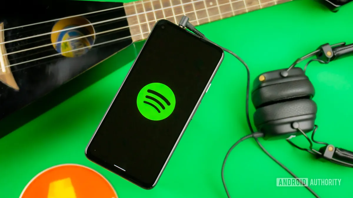 Teléfono con el logotipo de Spotify en pantalla sobre una superficie verde, junto a una guitarra y unos auriculares