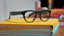Samsung smart glasses with dark green frame, transparent lenses and an integrated camera on the temple, over a stack of books