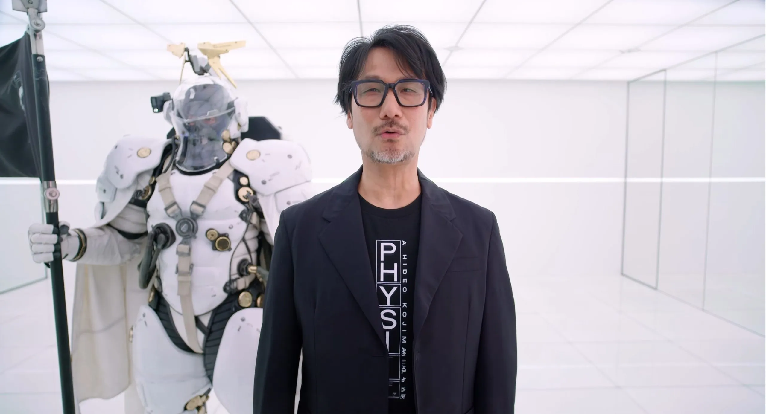 Hideo Kojima durante el anuncio de Physint, con una figura acorazada al fondo