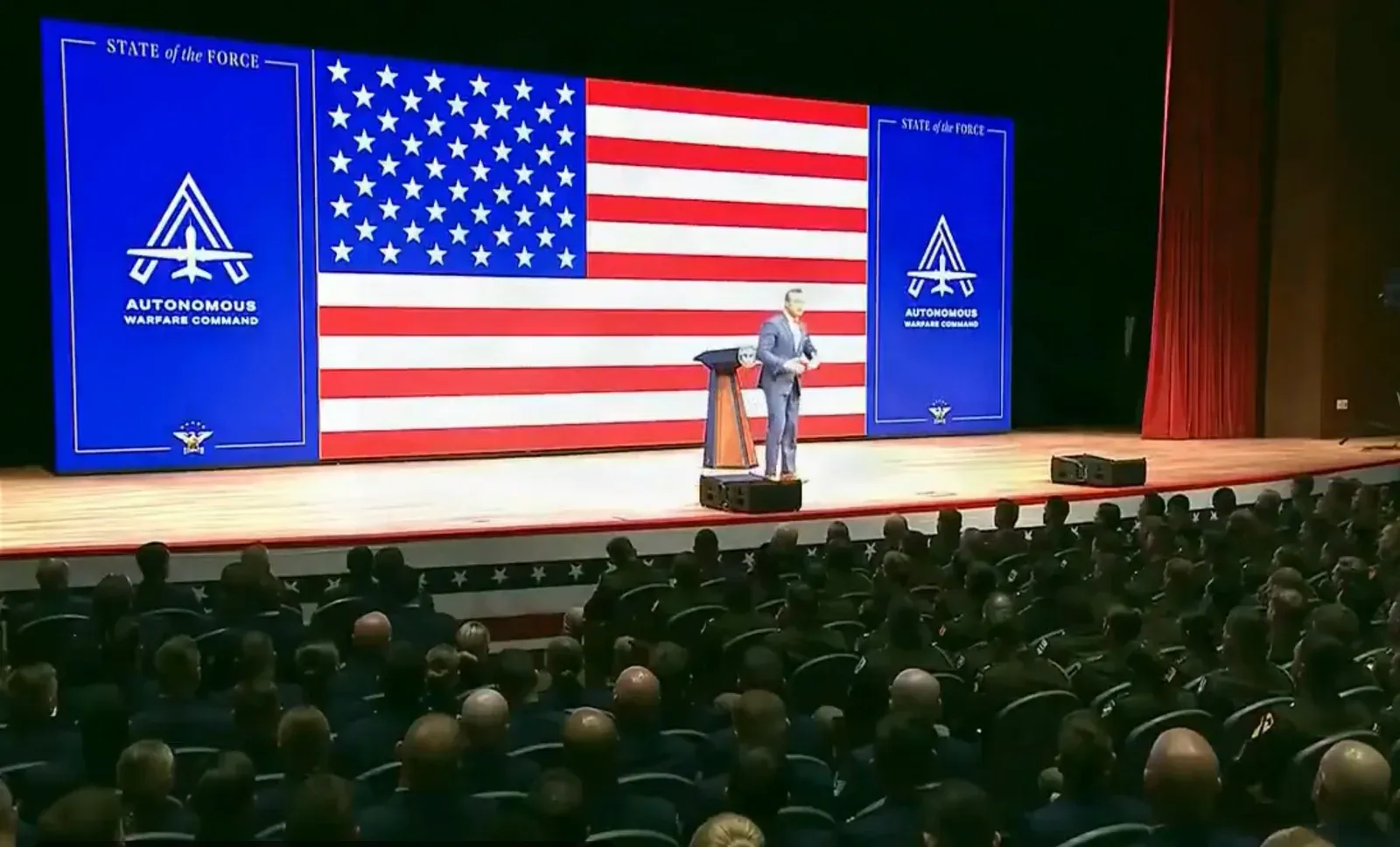 Wide view of the Quantico stage with a large US flag on screen, Hegseth at the lectern and rows of seated service members